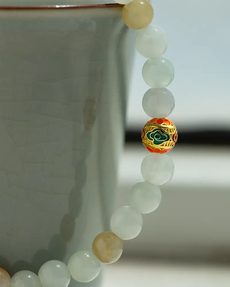 DSC_0595 拷贝 A close-up of a beaded bracelet with pale green and beige round beads, featuring a single ornate bead with red, gold, black, and green patterns, draped over the edge of a light gray ceramic cup.