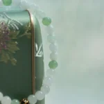 A beaded bracelet with alternating white and light green beads, plus a single black and gold bead, is draped over a green box with an embroidered floral design. Soft, muted background.