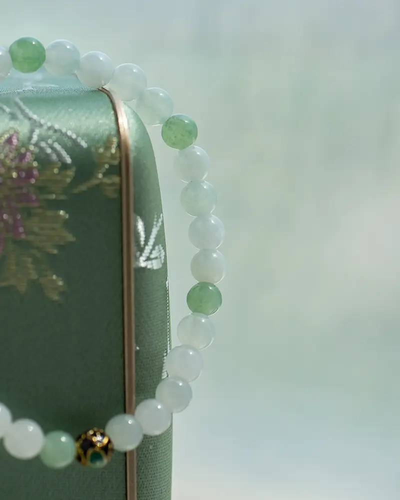 DSC_0600 拷贝 A beaded bracelet with alternating white and light green beads, plus a single black and gold bead, is draped over a green box with an embroidered floral design. Soft, muted background.