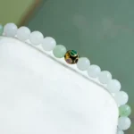 A beaded bracelet with mostly white and a few light green beads, featuring a single gold bead decorated with green and purple enamel, displayed on a white surface.