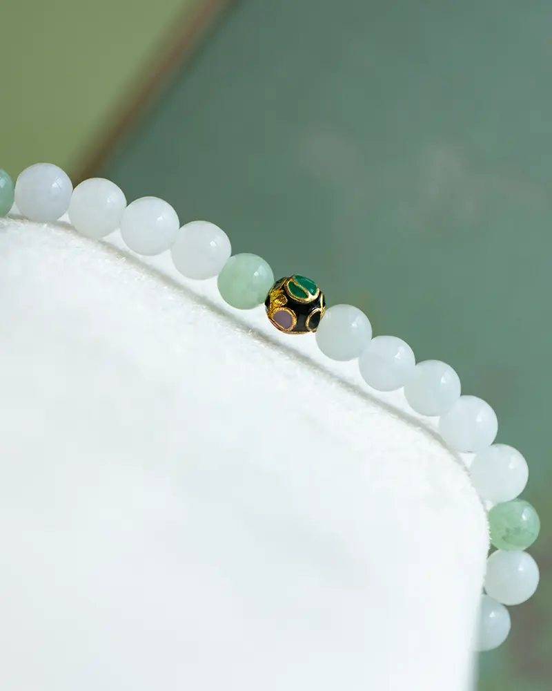 DSC_0606 拷贝 A beaded bracelet with mostly white and a few light green beads, featuring a single gold bead decorated with green and purple enamel, displayed on a white surface.