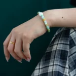 A hand rests on a knee, wearing a gold ring and a beaded bracelet with green, white, and yellow beads. The person is dressed in plaid pants against a dark green background.