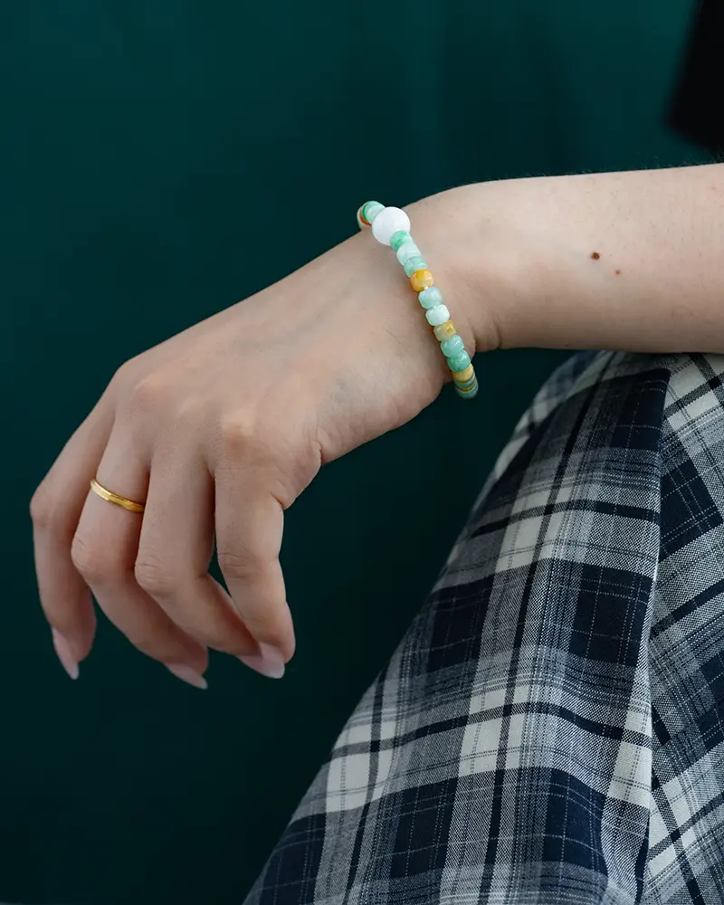 DSC_0611 拷贝 A hand rests on a knee, wearing a gold ring and a beaded bracelet with green, white, and yellow beads. The person is dressed in plaid pants against a dark green background.