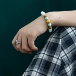 A hand wearing a green gemstone ring and a beaded bracelet with gold and green beads rests on a knee. The person is dressed in a black and white plaid skirt with a dark green background behind them.