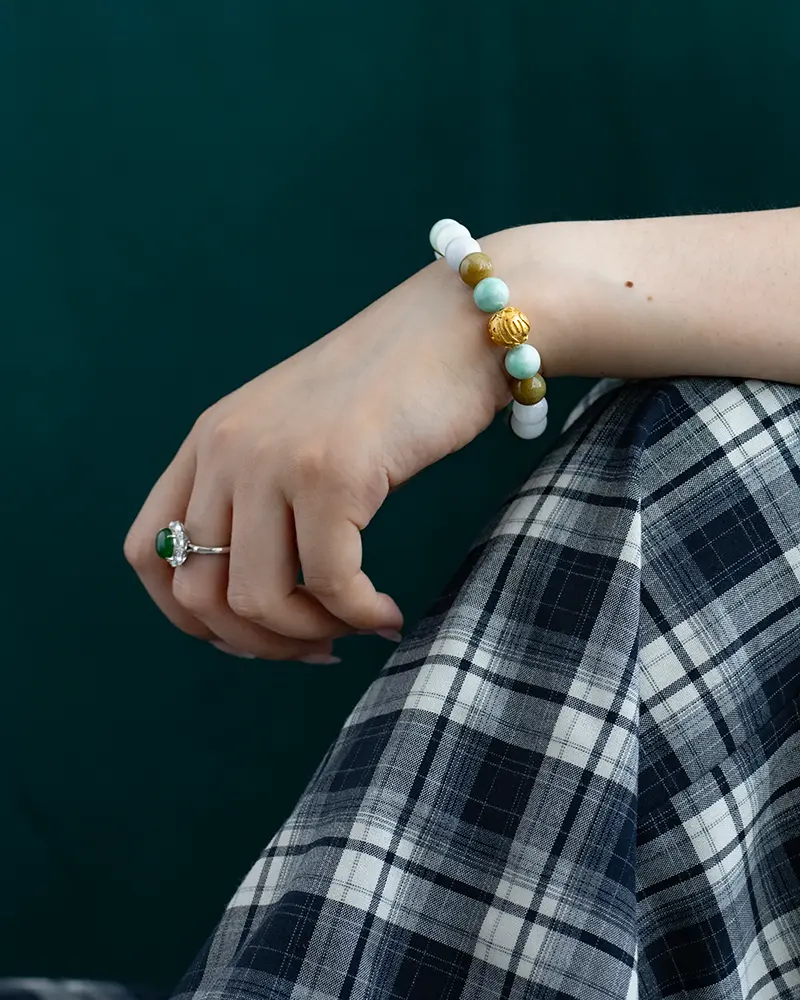 DSC_0615 拷贝 A hand wearing a green gemstone ring and a beaded bracelet with gold and green beads rests on a knee. The person is dressed in a black and white plaid skirt with a dark green background behind them.