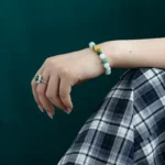 A hand wearing a jade beaded bracelet with gold accents and a silver ring with a green stone, resting on a plaid skirt against a dark green background.