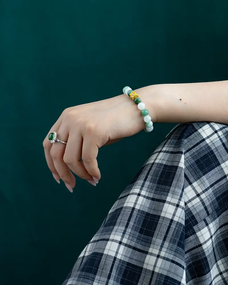 DSC_0616 拷贝 A hand wearing a jade beaded bracelet with gold accents and a silver ring with a green stone, resting on a plaid skirt against a dark green background.