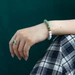 A hand wearing a green gemstone ring and a beaded bracelet with green and white beads rests on a plaid-patterned fabric, with a dark green background.