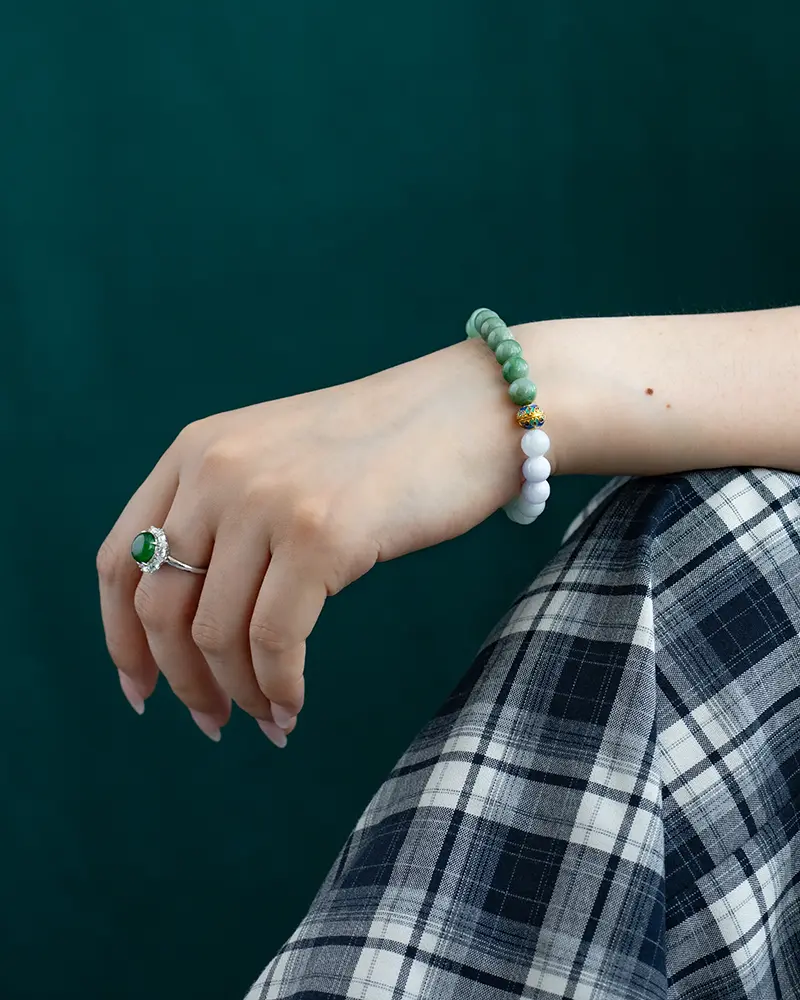 DSC_0618 拷贝 A hand wearing a green gemstone ring and a beaded bracelet with green and white beads rests on a plaid-patterned fabric, with a dark green background.