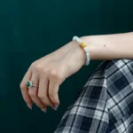A hand wearing a beaded bracelet with a gold accent and an emerald ring rests on a knee. The person is dressed in a black and white plaid skirt, and the background is dark green.