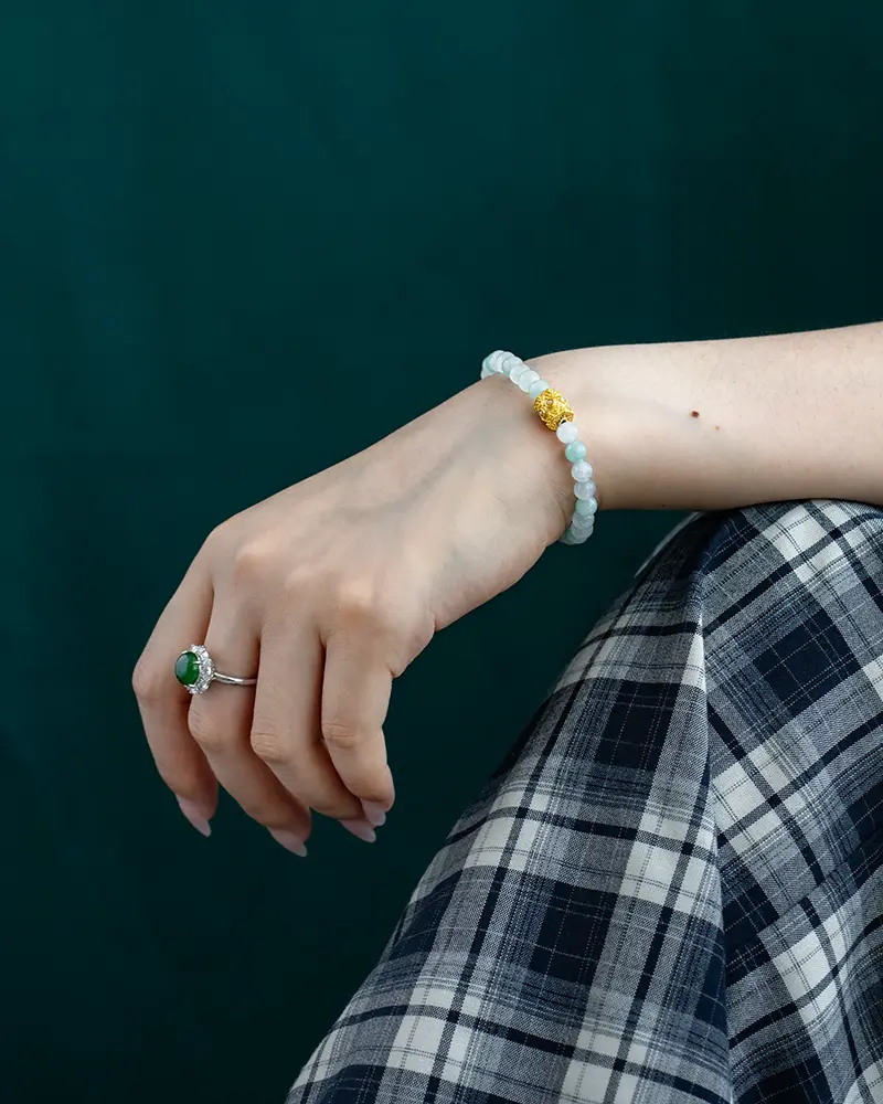 DSC_0620 拷贝 A hand wearing a beaded bracelet with a gold accent and an emerald ring rests on a knee. The person is dressed in a black and white plaid skirt, and the background is dark green.