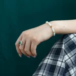 A hand wearing a green gemstone ring and a beaded bracelet rests on a plaid-patterned skirt in front of a dark green background.
