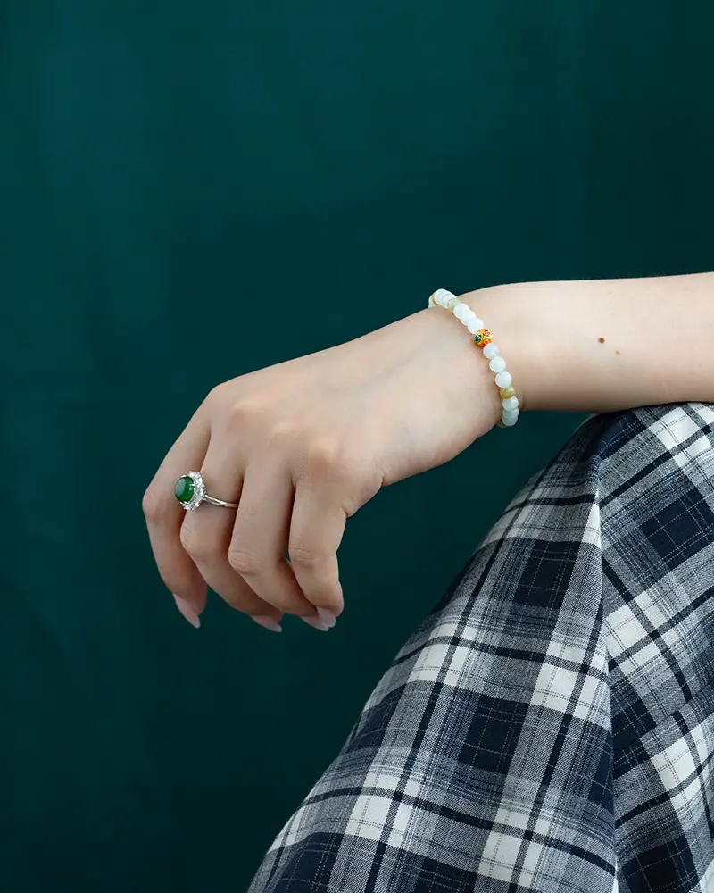 DSC_0624 拷贝 A hand wearing a green gemstone ring and a beaded bracelet rests on a plaid-patterned skirt in front of a dark green background.