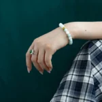 A hand with a silver ring and a beaded bracelet rests on a plaid-patterned skirt against a dark green background.