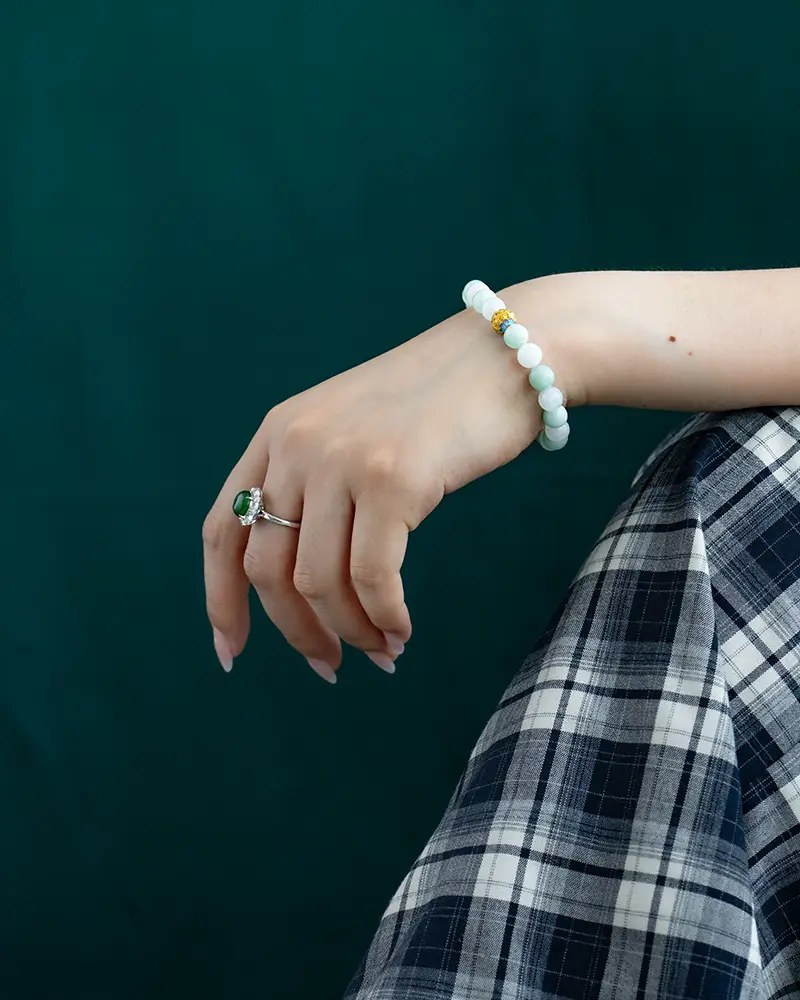 A hand with a silver ring and a beaded bracelet rests on a plaid-patterned skirt against a dark green background.