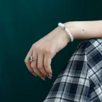 A hand resting on a lap wearing a green gemstone ring and a white beaded bracelet with a blue accent, against a dark green background. The person is dressed in a plaid-patterned skirt.