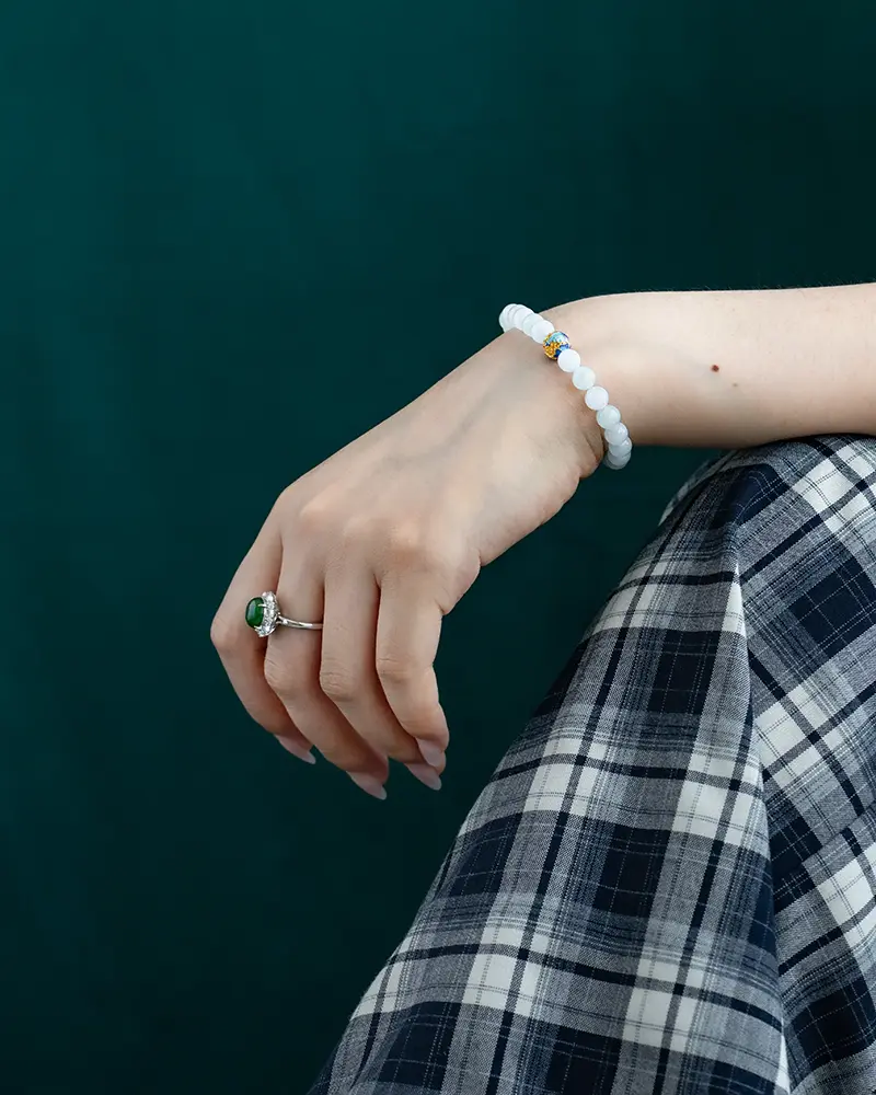 DSC_0627 拷贝 A hand resting on a lap wearing a green gemstone ring and a white beaded bracelet with a blue accent, against a dark green background. The person is dressed in a plaid-patterned skirt.