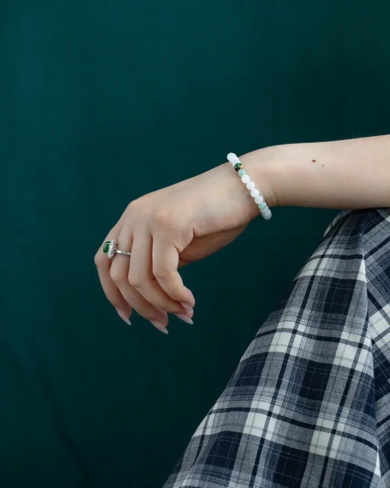 A hand adorned with the Light Green and White Jadeite Bead Bracelet—featuring spherical beads and an enamel-style accent—rests on a lap draped in a blue and white plaid skirt, set against a dark green background.