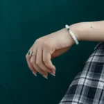 A hand with pale nail polish wears a beaded white bracelet and a silver ring with a green gemstone, resting against a dark green background and a plaid skirt.