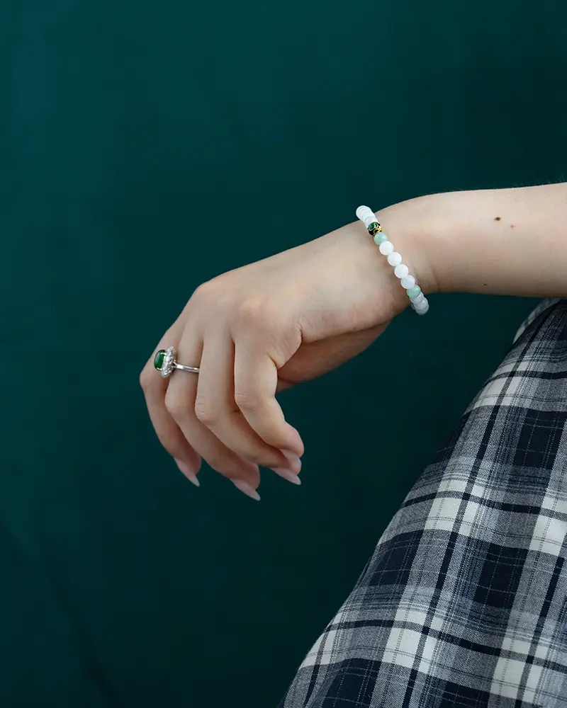 DSC_0629 拷贝 A hand with pale nail polish wears a beaded white bracelet and a silver ring with a green gemstone, resting against a dark green background and a plaid skirt.