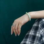 A persons hand with manicured nails rests on their knee, wearing a green gemstone ring and a light green beaded bracelet. The person is dressed in a blue and white plaid skirt against a dark green background.