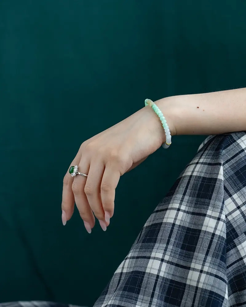 A persons hand with manicured nails rests on their knee, wearing a green gemstone ring and a light green beaded bracelet. The person is dressed in a blue and white plaid skirt against a dark green background.