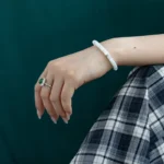 A hand wearing a jade bracelet and a silver ring with a green stone rests on a lap covered in blue and white plaid fabric, with a dark green background.