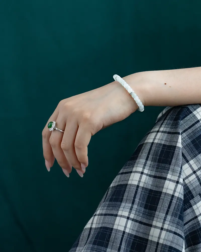 DSC_0634 拷贝 A hand wearing a jade bracelet and a silver ring with a green stone rests on a lap covered in blue and white plaid fabric, with a dark green background.