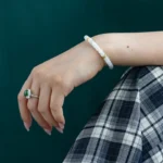 A hand wearing a jade and gold bracelet and a green gemstone ring rests on a plaid-patterned skirt, with a dark green background.