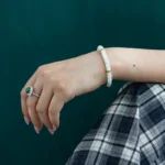 A hand wearing a white beaded bracelet and a silver ring with a green stone rests on a knee, which is covered by black-and-white plaid fabric, against a dark green background.