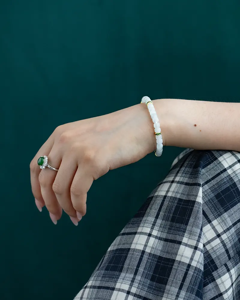 DSC_0637 拷贝 A hand wearing a white beaded bracelet and a silver ring with a green stone rests on a knee, which is covered by black-and-white plaid fabric, against a dark green background.