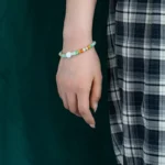 A persons hand with a beaded bracelet hangs by their side. They are wearing a black and white plaid skirt, and the background is dark green.