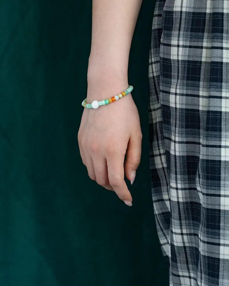 DSC_0644 拷贝 A persons hand with a beaded bracelet hangs by their side. They are wearing a black and white plaid skirt, and the background is dark green.