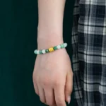 A person wearing a green beaded bracelet with a central gold bead rests their hand by their side. The person is dressed in a black and white plaid skirt, and the background is dark green.