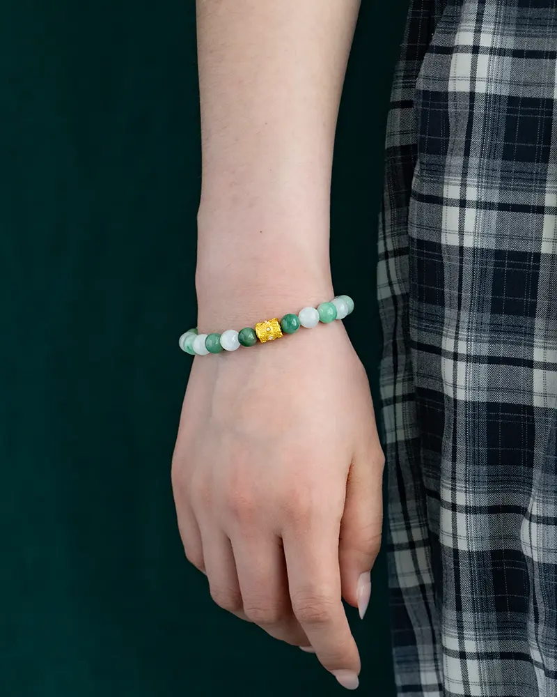 DSC_0646 拷贝 A person wearing a green beaded bracelet with a central gold bead rests their hand by their side. The person is dressed in a black and white plaid skirt, and the background is dark green.