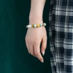 A person wearing a plaid skirt and a beaded bracelet with green, white, and gold beads stands against a dark green background.