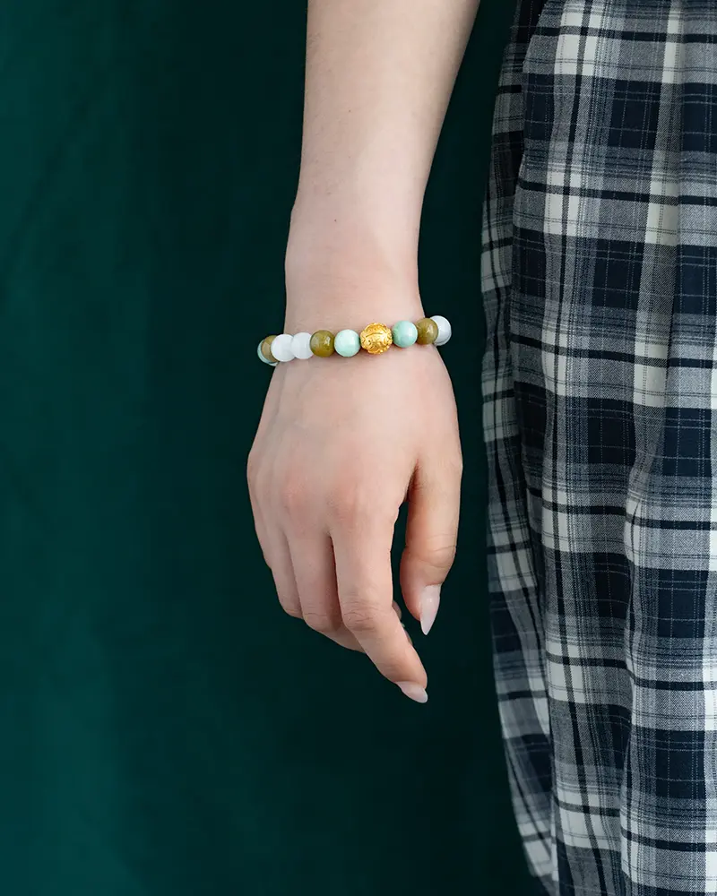 DSC_0647 拷贝 A person wearing a plaid skirt and a beaded bracelet with green, white, and gold beads stands against a dark green background.