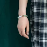 A person wearing a black-and-white plaid skirt and a beaded bracelet with green and white beads stands against a dark green background. Only the lower arm and hand are visible.