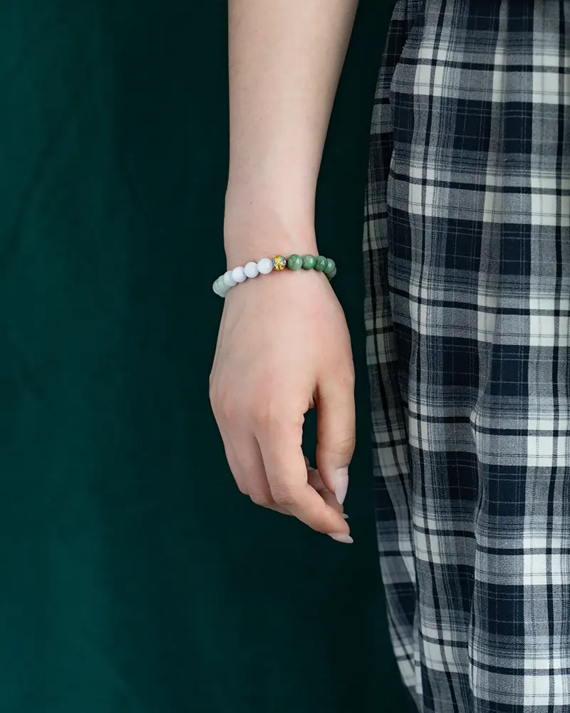 DSC_0648 拷贝 A person wearing a black-and-white plaid skirt and a beaded bracelet with green and white beads stands against a dark green background. Only the lower arm and hand are visible.