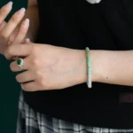 A person wearing a black top and plaid skirt displays a green stone ring on one hand and a beaded green and white bracelet on the other wrist. Only the hands and part of the torso are visible.