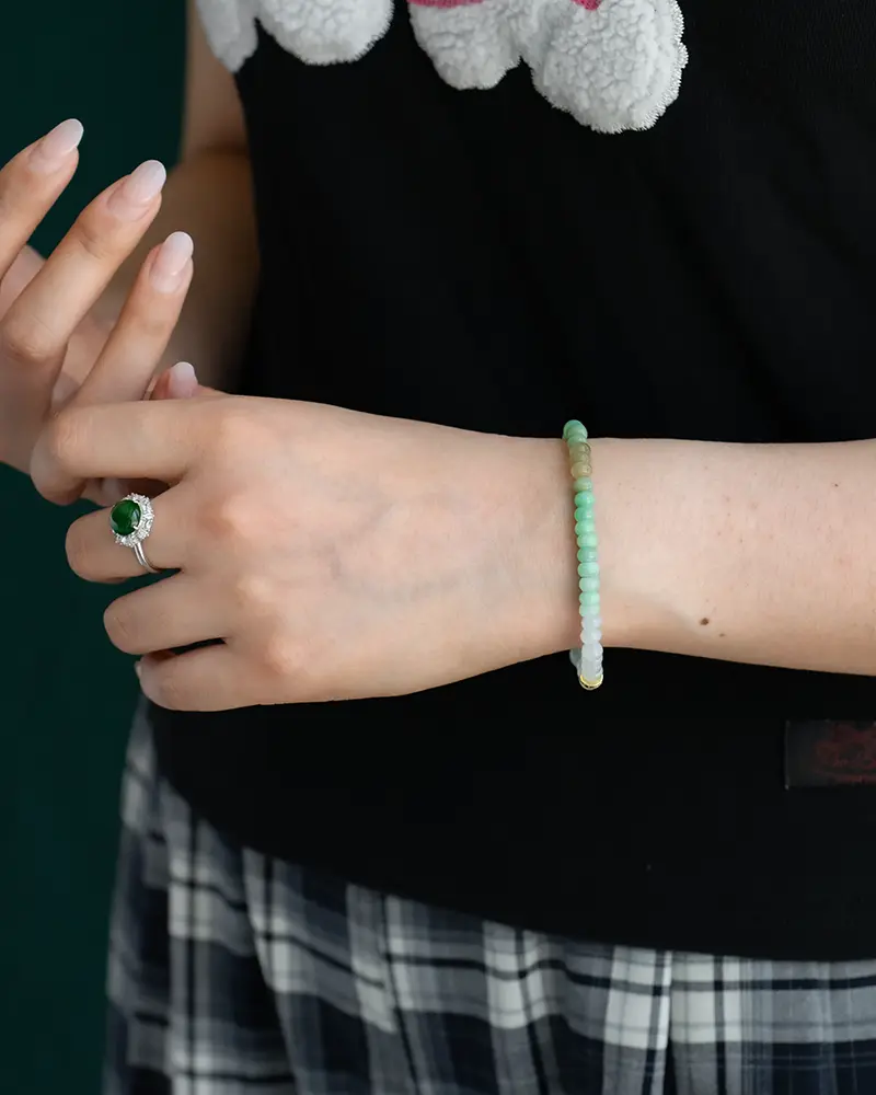 A person wearing a black top and plaid skirt displays a green stone ring on one hand and a beaded green and white bracelet on the other wrist. Only the hands and part of the torso are visible.