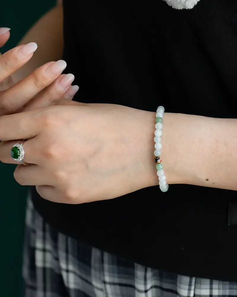 DSC_0652 拷贝 A person wearing a beaded bracelet with white and green beads on their wrist, and a silver ring with a large green stone on their finger. Their nails are manicured, and they are dressed in a black top and plaid skirt.