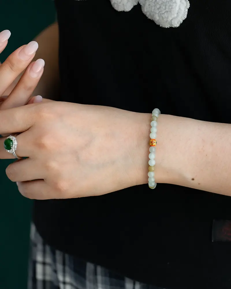 DSC_0653 拷贝 A close-up of a person’s hand wearing a beaded bracelet with light green and one gold bead, and a silver ring with a green gemstone. The person is dressed in a black top and plaid skirt.