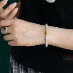 A person wearing a beaded bracelet with a gold accent on their wrist, a silver ring with a green gemstone on a finger, a black shirt, and a plaid skirt.
