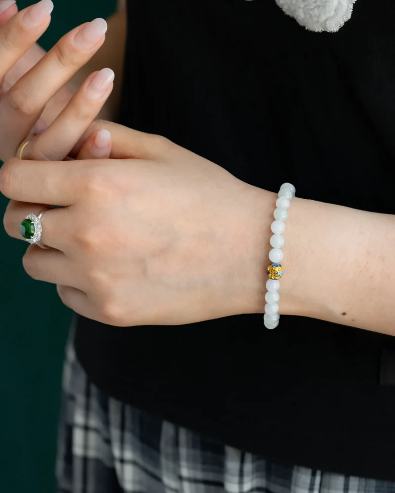 A person wearing the White and Light Green Jadeite Bead Bracelet featuring spherical beads with a blue accent detail, along with a silver ring set with a green stone, dressed in a black top and plaid skirt.