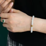 A person wearing a beaded bracelet with a single gold accent and a silver ring with a green gemstone, dressed in a black top and plaid skirt, is shown adjusting the bracelet on their wrist.