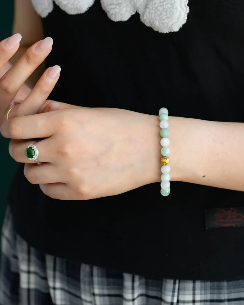 A person wearing a Light Green and White Jadeite Bead Bracelet with spherical beads and a gold-tone accent, paired with a green gemstone ring, black top, plaid skirt, and showing manicured nails.
