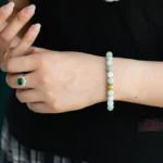 A person wearing a jade bead bracelet with a gold accent on their wrist. They also wear a green gemstone ring and have manicured nails. The person is dressed in a black top and plaid skirt.