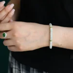 A person wearing a beaded bracelet with gold accents on their wrist and a silver ring with a green stone on their finger, dressed in a black top and plaid skirt.
