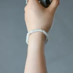 A hand wearing a delicate white beaded bracelet holds a small cup. The person also has a green gemstone ring on their index finger. The background is plain and gray.